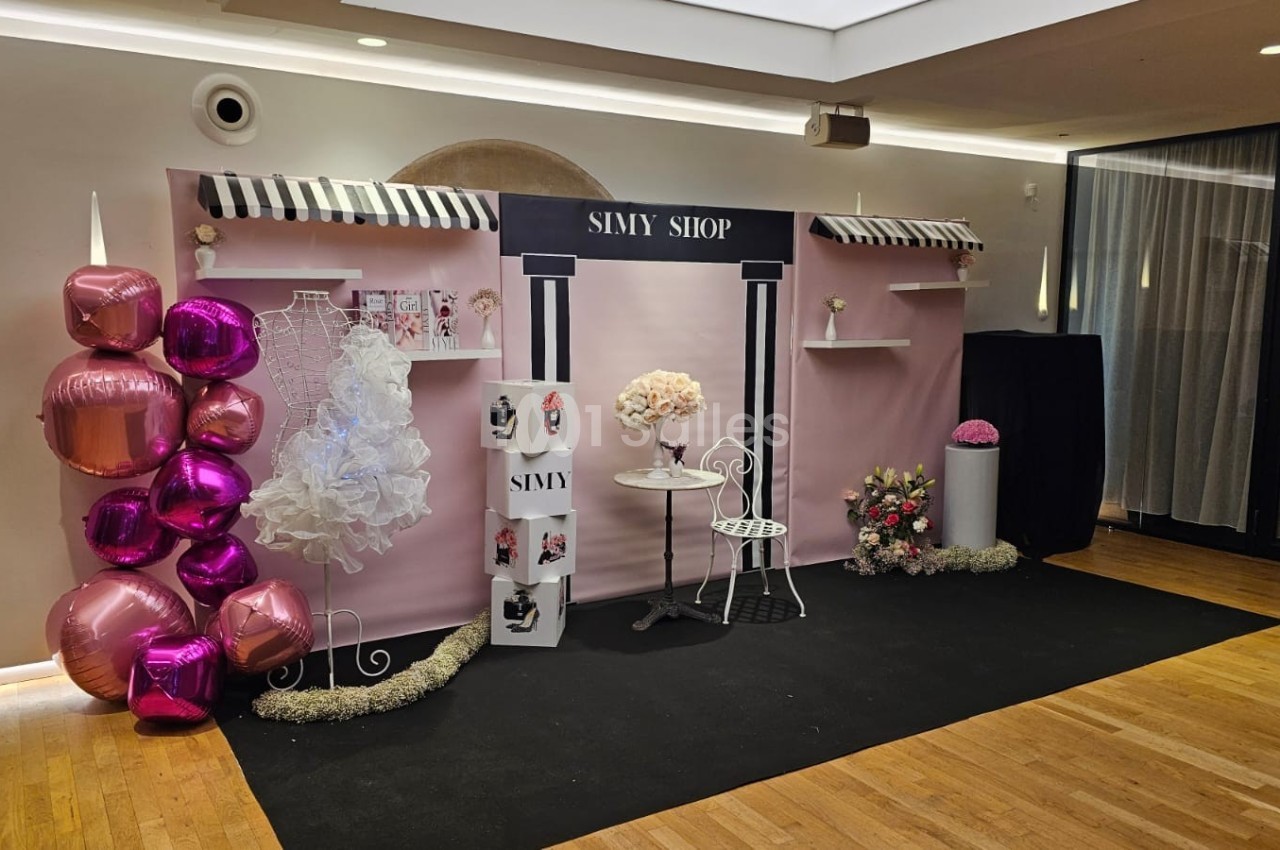 Décor rose avec ballons, présentoirs, robe blanche, table et chaise, évoquant une boutique ou un espace d'exposition.