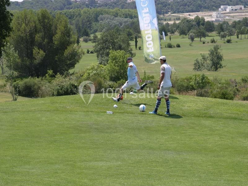 Deux enfants jouent au football sur un terrain herbeux face à un paysage vallonné boisé.