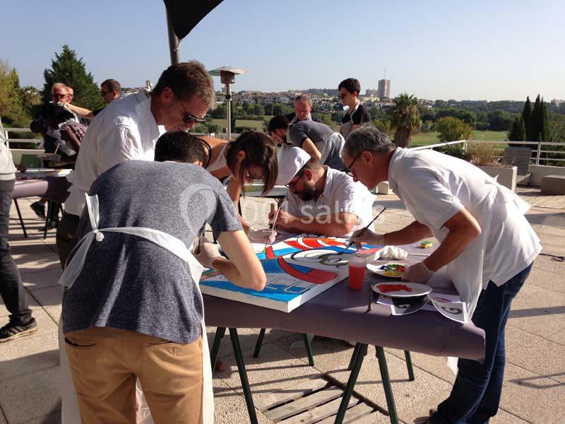 Groupe de personnes réunies autour d'une table en terrasse ensoleillée, vue sur la campagne environnante.