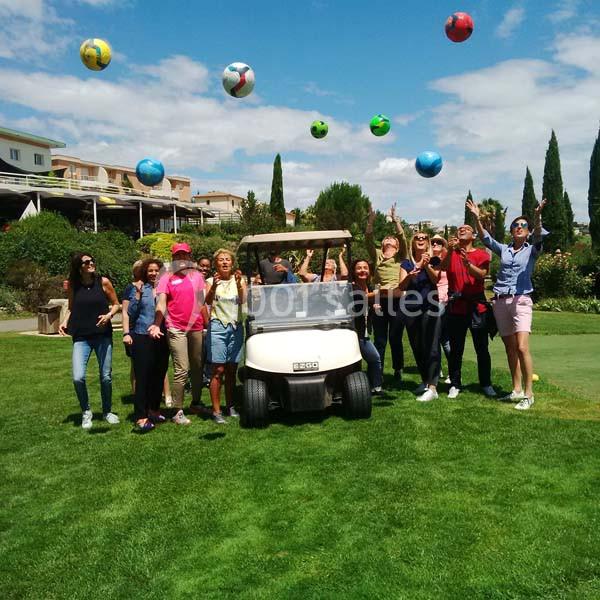 Groupe de personnes autour d'un buggy sur pelouse ensoleillée, balles colorées en l'air, bâtiments et cyprès en arrière-plan.