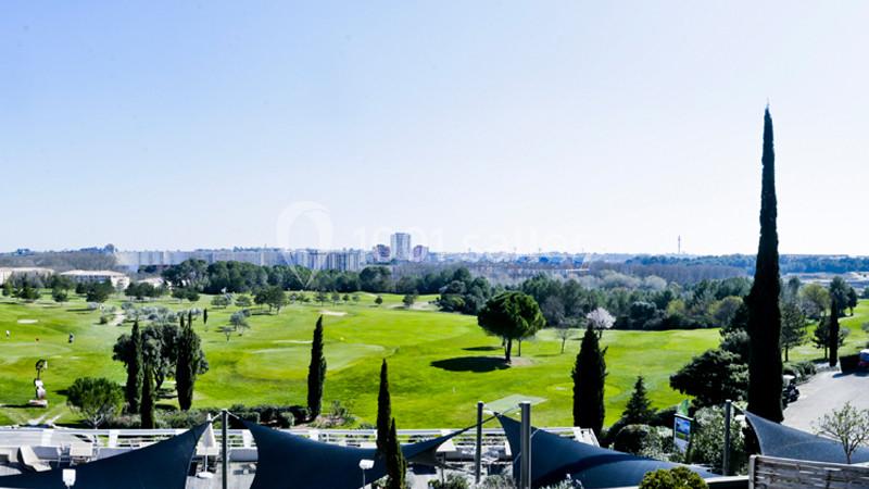 Vue depuis une terrasse sur un golf verdoyant avec cyprès, skyline urbaine en arrière-plan et ciel dégagé.