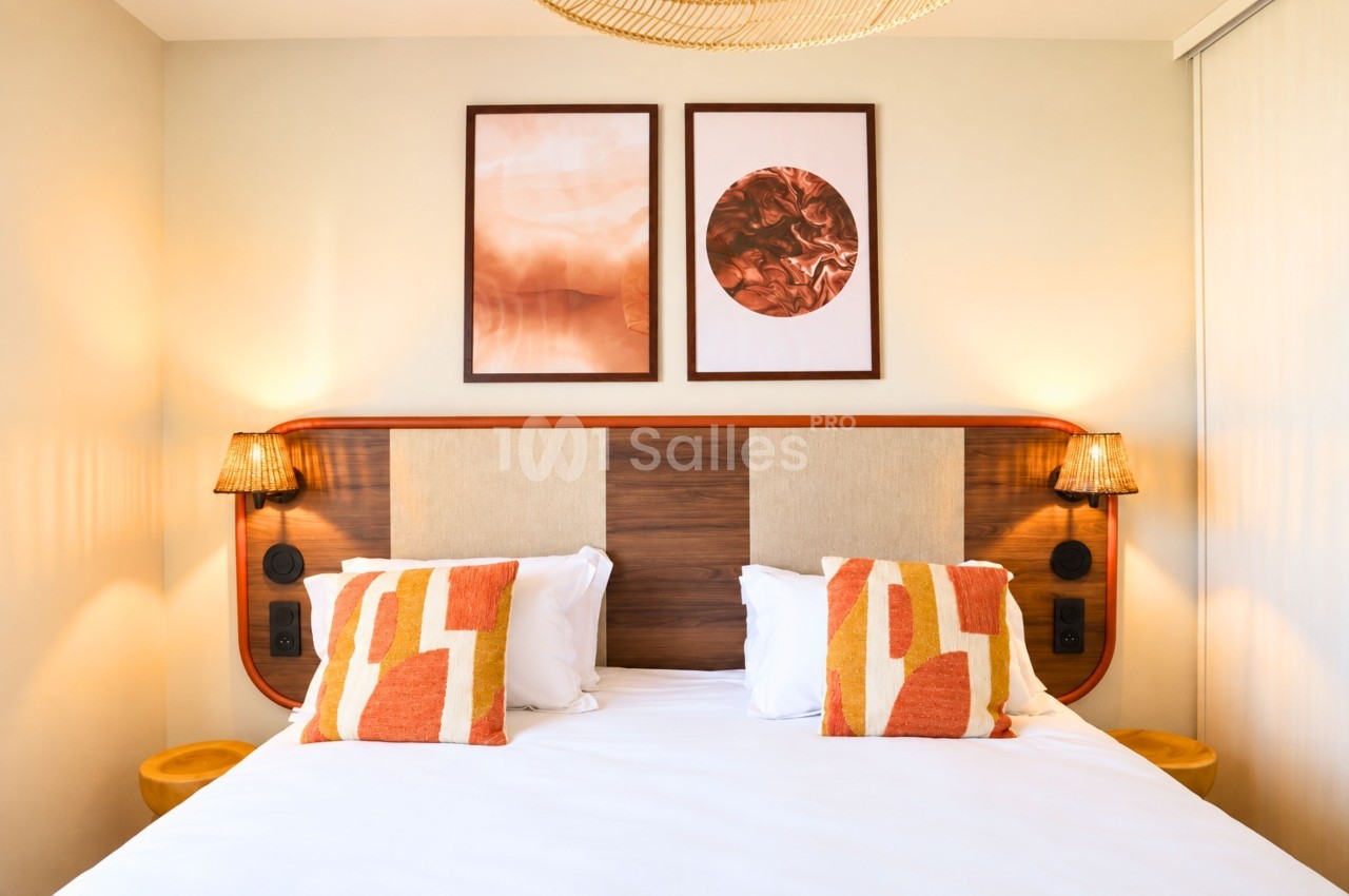Chambre double avec lit en bois, coussins orange et blanc, deux lampes de chevet, deux tableaux muraux aux tons chauds.