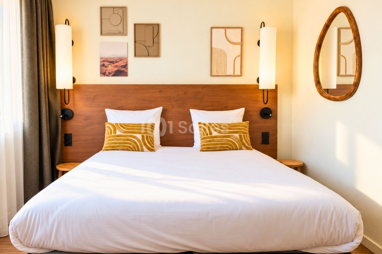 Chambre d'hôtel avec lit double aux draps blancs, tête de lit en bois clair et lampes murales intégrées.