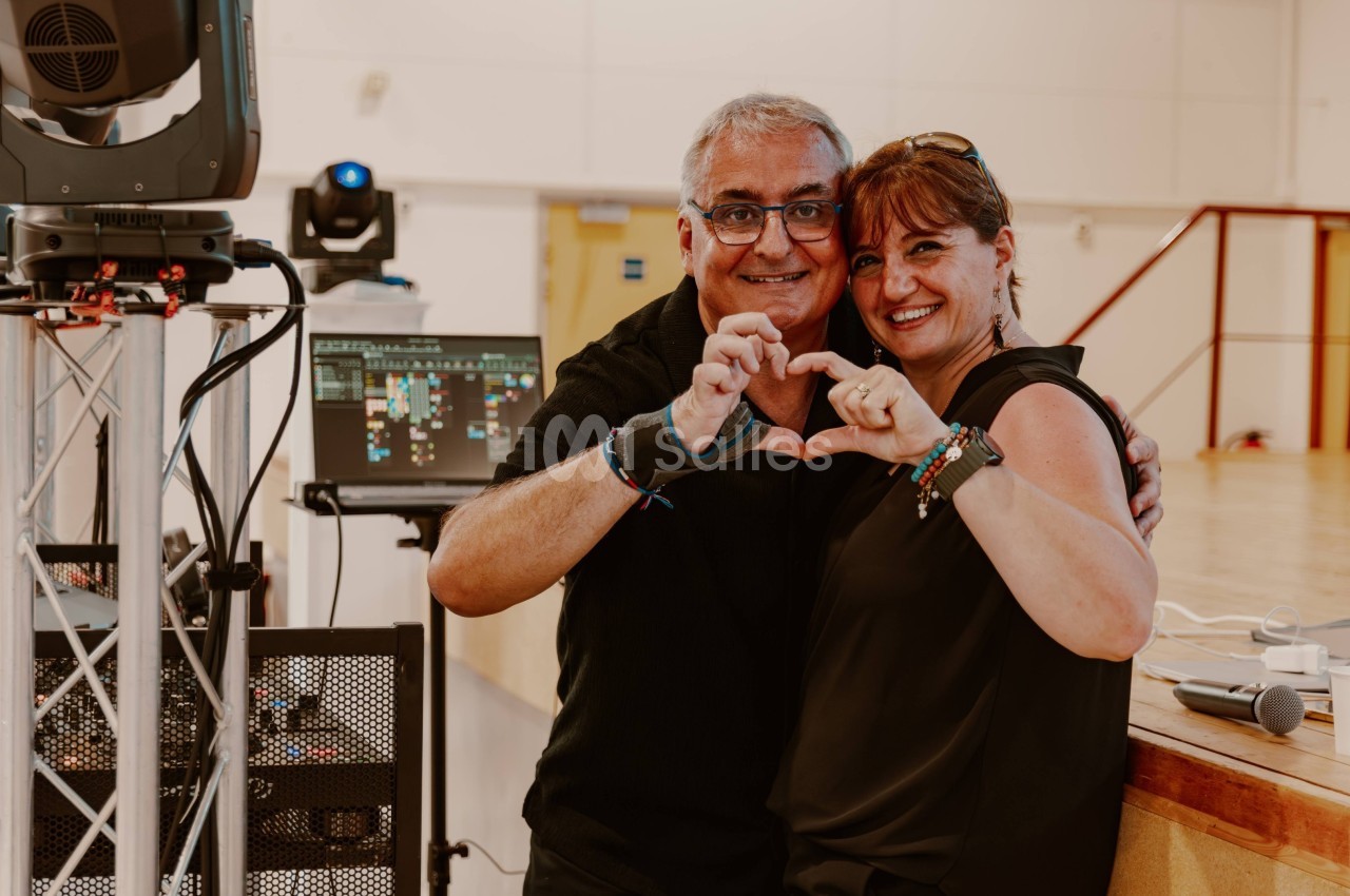 Un homme et une femme souriants forment un cœur avec leurs mains dans une salle équipée de matériel audiovisuel.