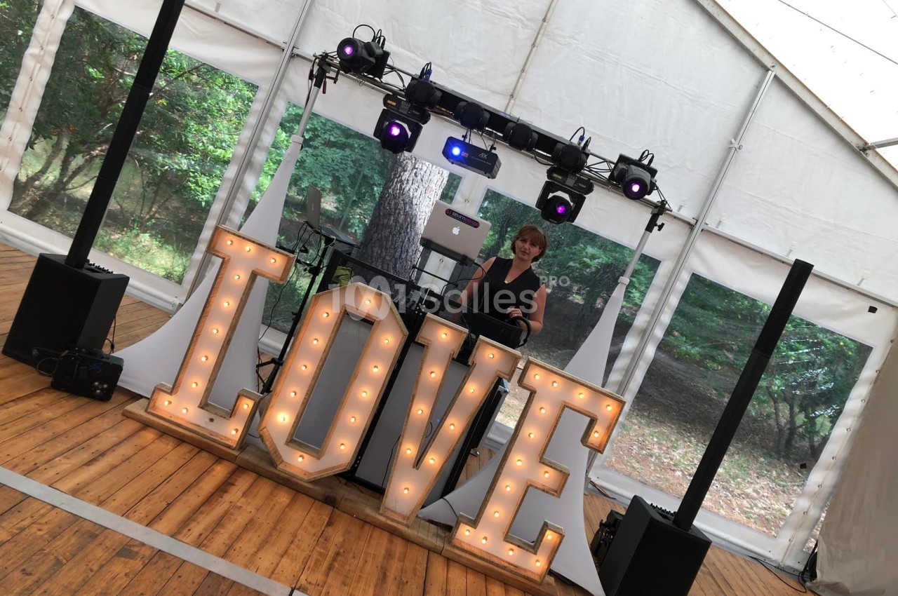 Un DJ mixe derrière une table décorée de grandes lettres lumineuses formant le mot ’LOVE’ dans une tente ouverte.