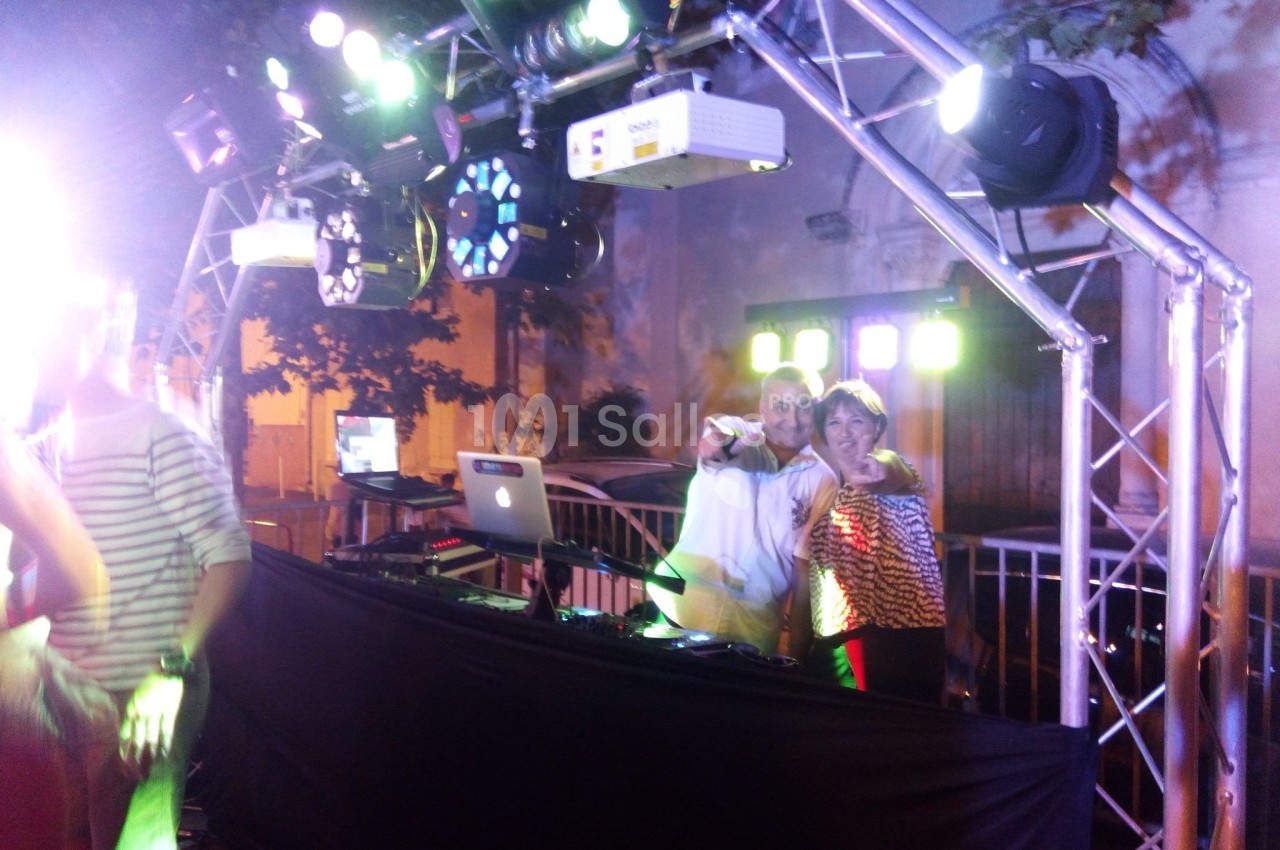 Un DJ et une femme posent devant une table de mixage sous une structure lumineuse lors d'un événement en extérieur.
