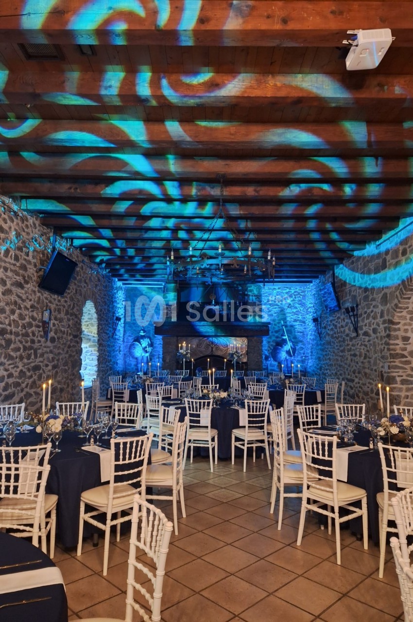 Salle en pierre décorée pour un événement, avec tables dressées, chaises blanches et éclairage bleu sur le plafond.