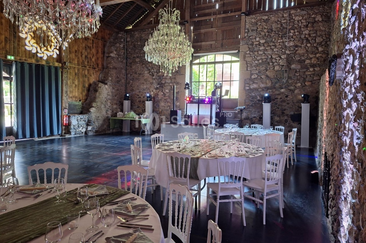 Salle de réception rustique avec murs en pierre, tables dressées, chaises blanches et lustres suspendus.