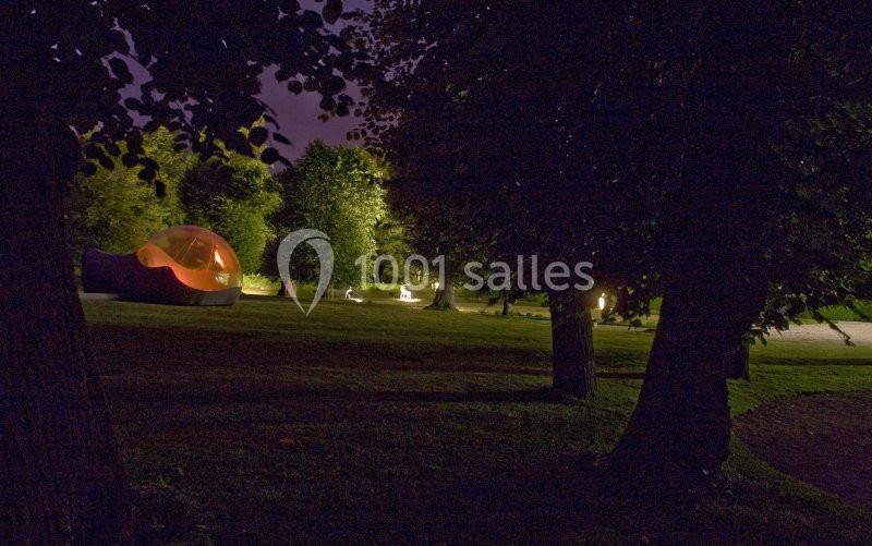 Un parc de nuit avec des arbres sombres, une structure lumineuse orange et des chemins faiblement éclairés.
