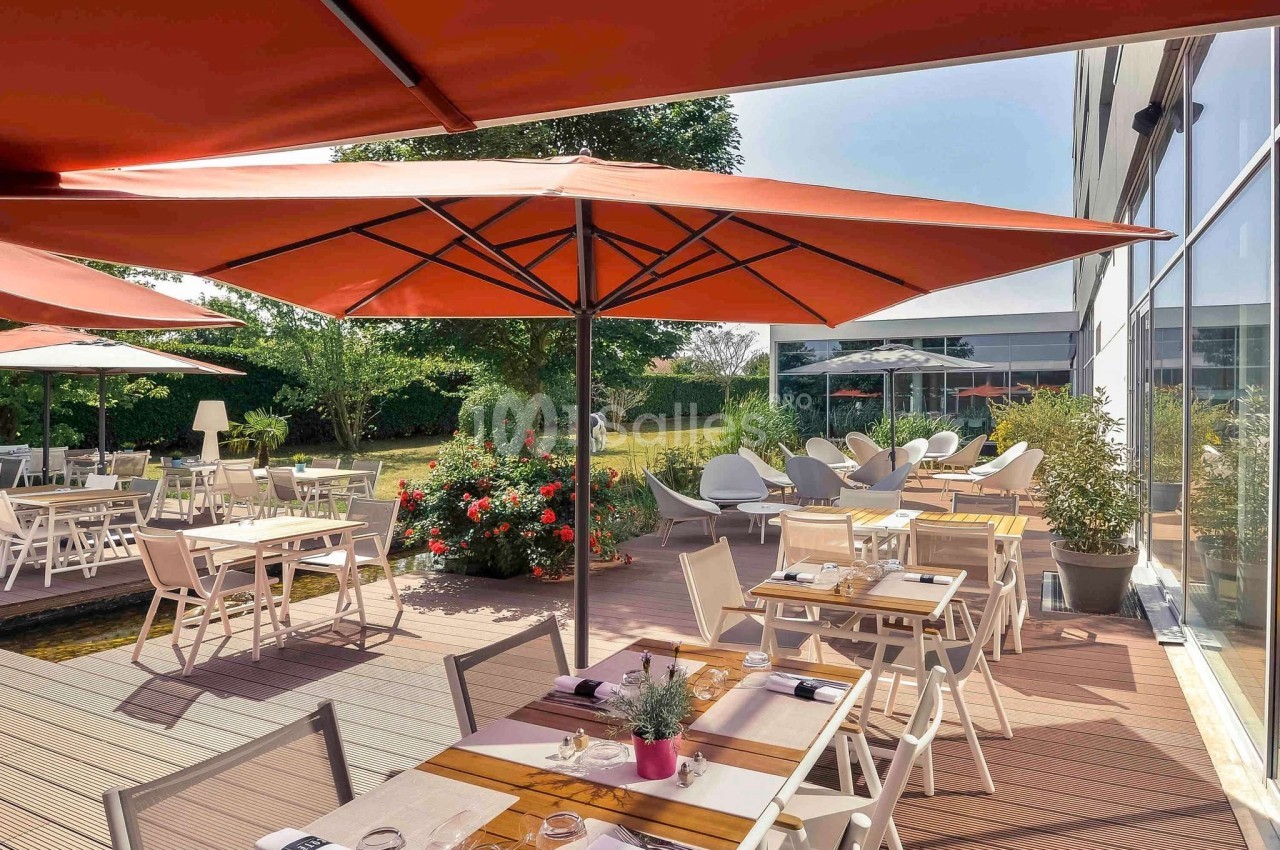 Terrasse d'un restaurant avec tables dressées, parasols rouges et vue sur un jardin fleuri.