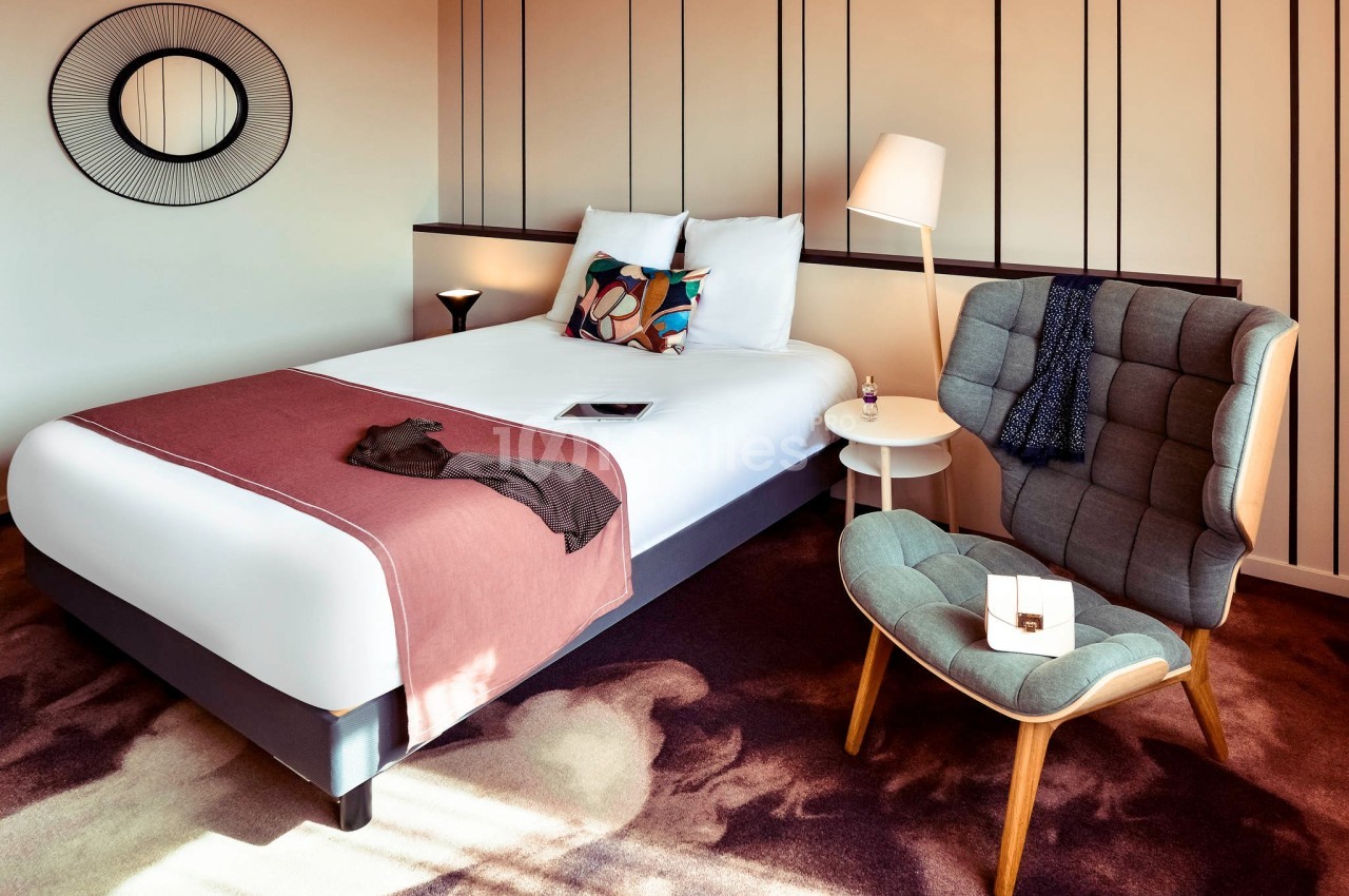Chambre d'hôtel moderne avec lit double, fauteuil, lampe, table d'appoint et décoration murale minimaliste.