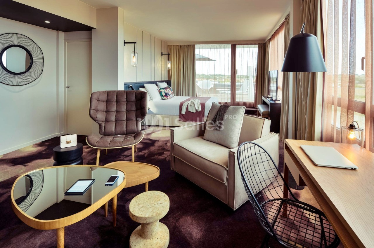 Chambre d'hôtel moderne avec lit, coin salon, bureau, grandes fenêtres et décoration contemporaine.