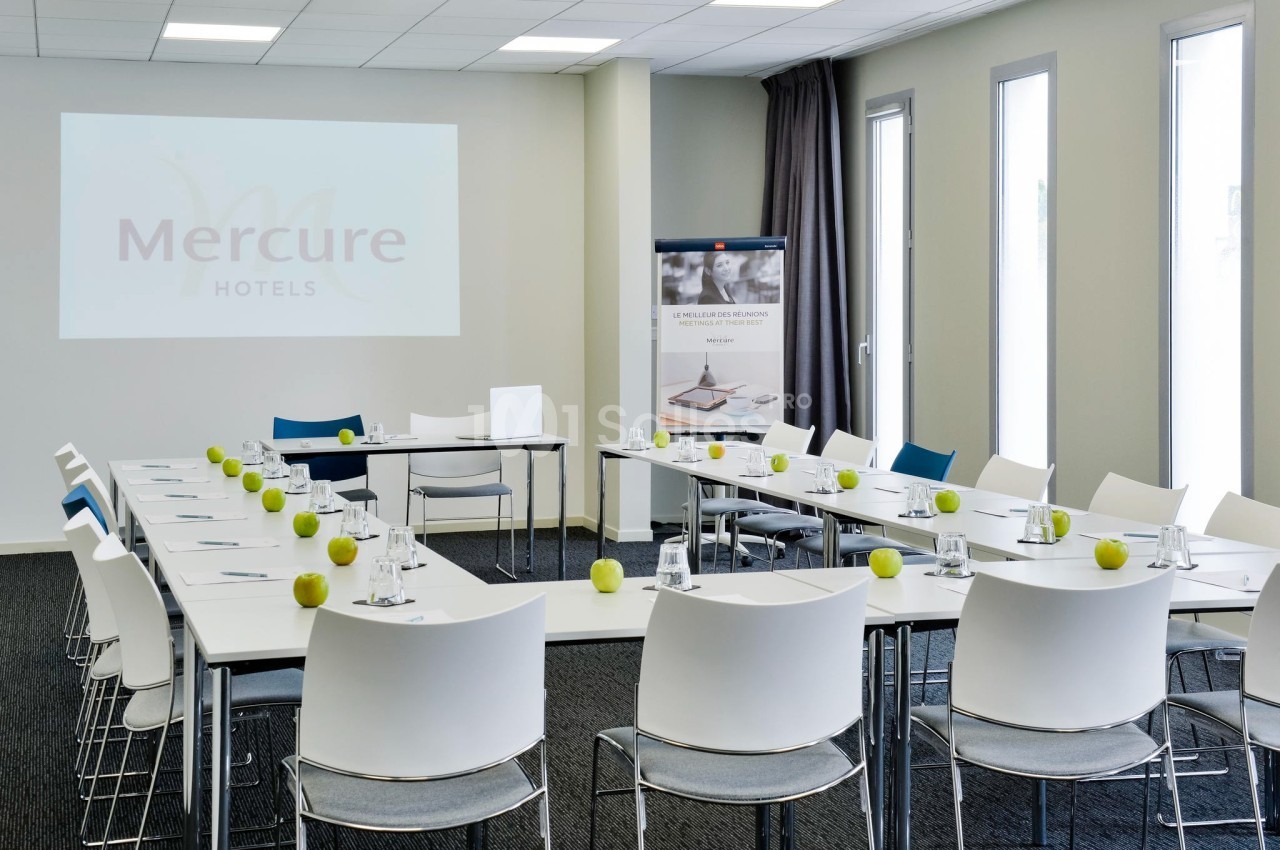 Salle de réunion lumineuse avec tables disposées en U, chaises blanches, pommes vertes et écran de projection.