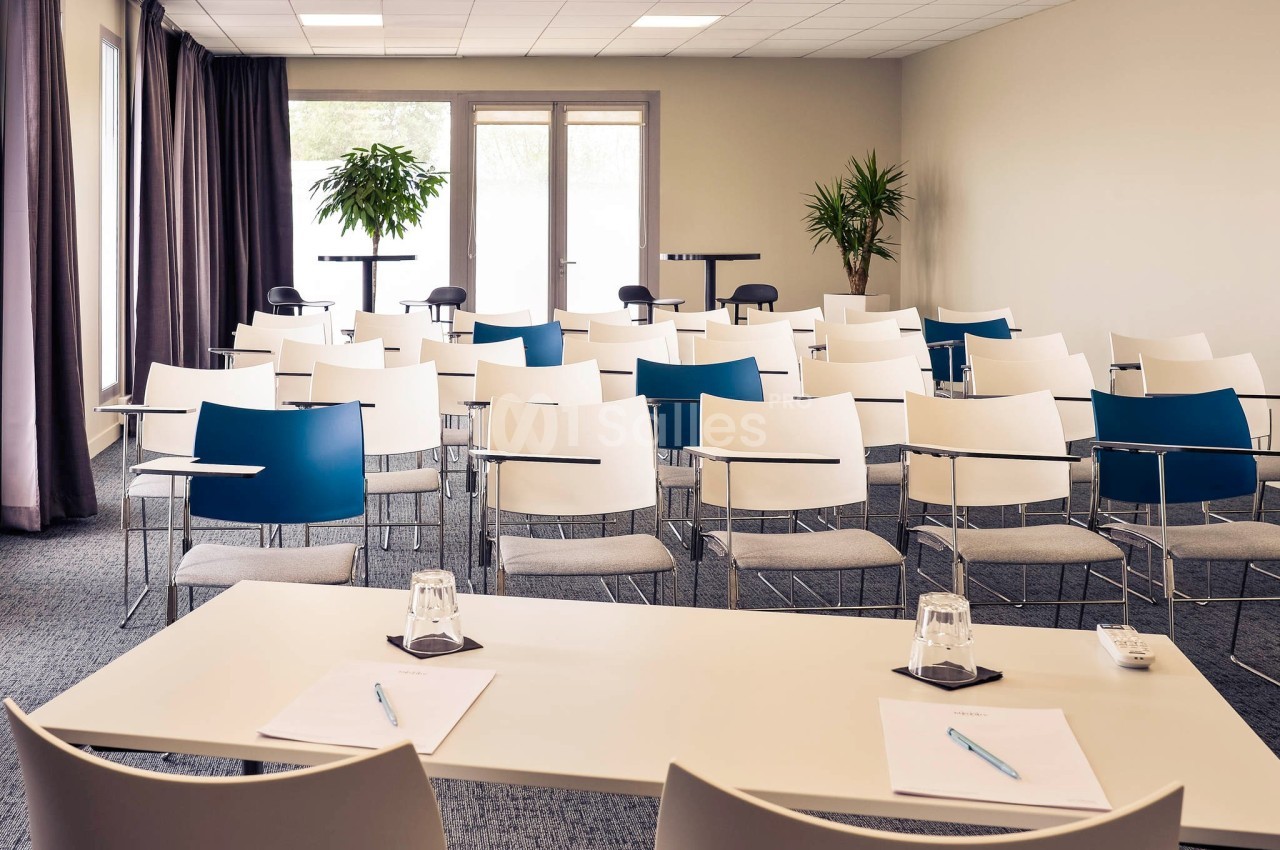 Salle de réunion lumineuse avec des rangées de chaises blanches et bleues, vue depuis une table équipée de stylos et verres.
