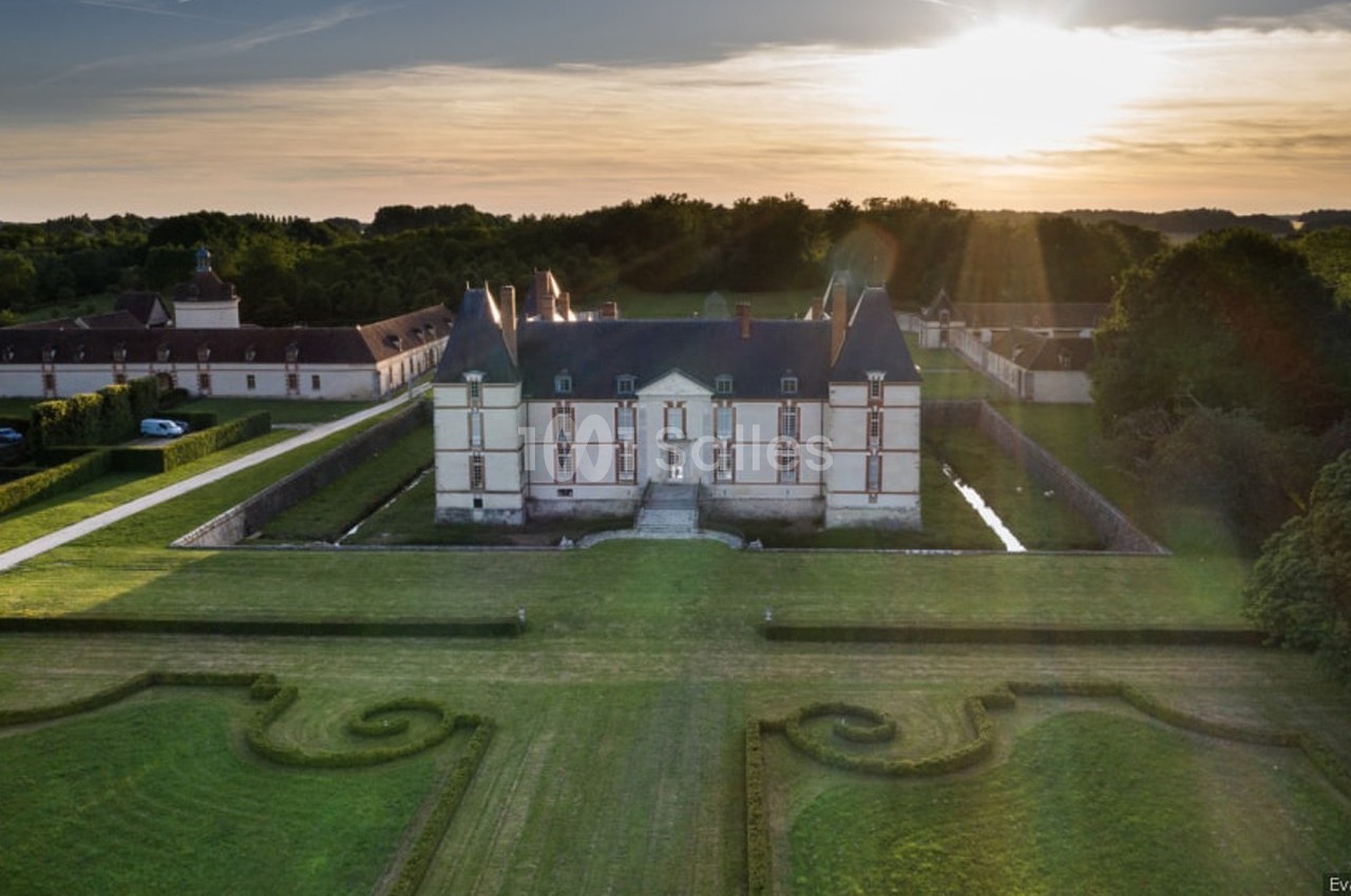 Vue aérienne d'un château entouré de jardins symétriques et de douves, éclairé par un coucher de soleil.