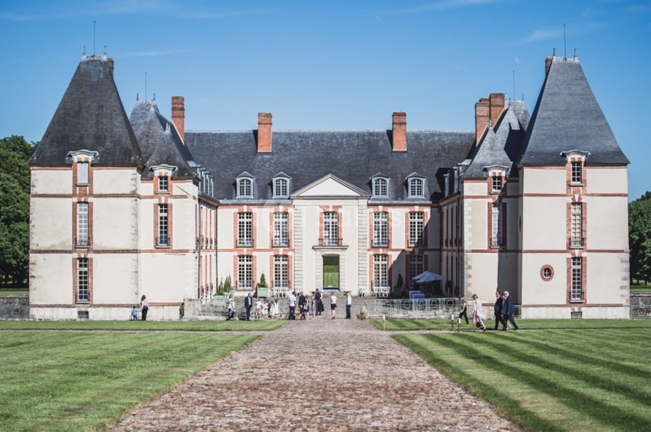 Façade d'un château avec deux tours, une allée pavée au premier plan et des visiteurs dans la cour.