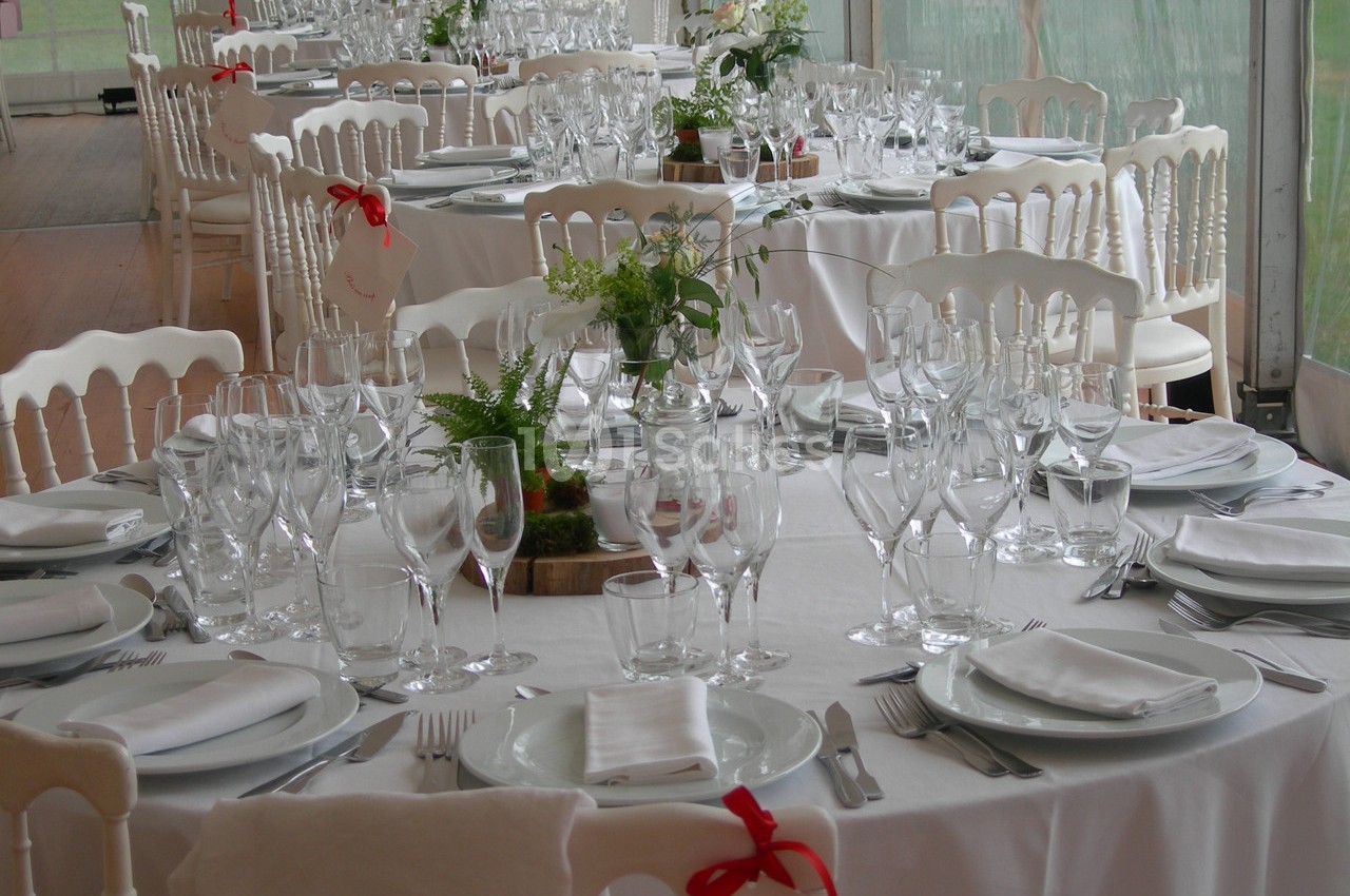 Tables rondes élégamment dressées avec nappes blanches, verres, couverts et décorations florales dans une salle lumineuse.