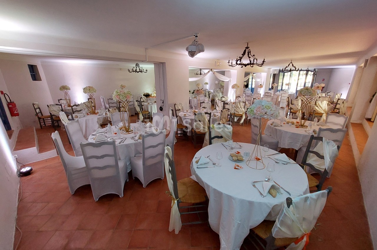 Salle de réception décorée avec des tables rondes, nappes blanches, chaises habillées et centres de table fleuris.
