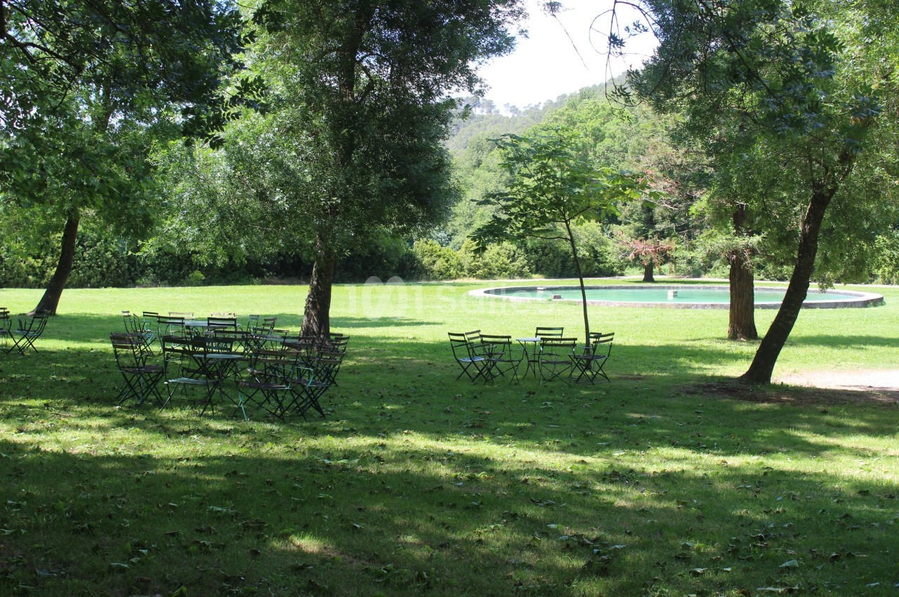 Chaises et tables en métal disposées sur une pelouse ombragée, avec une fontaine entourée de verdure en arrière-plan.