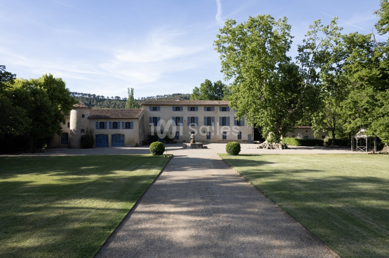 Allée gravillonnée menant à une grande bâtisse en pierre entourée de pelouses et d'arbres sous un ciel dégagé.