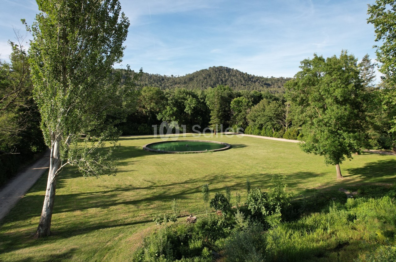 Vue d'un grand espace vert avec une pelouse, un bassin circulaire au centre et des arbres en arrière-plan.
