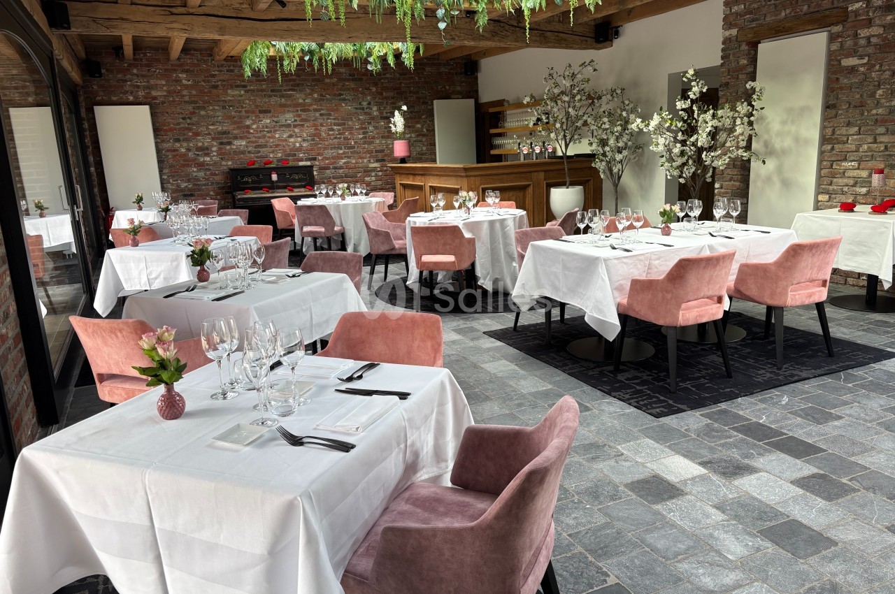 Salle de restaurant lumineuse avec tables dressées, chaises roses, murs en briques et plafond en bois décoré de plantes.