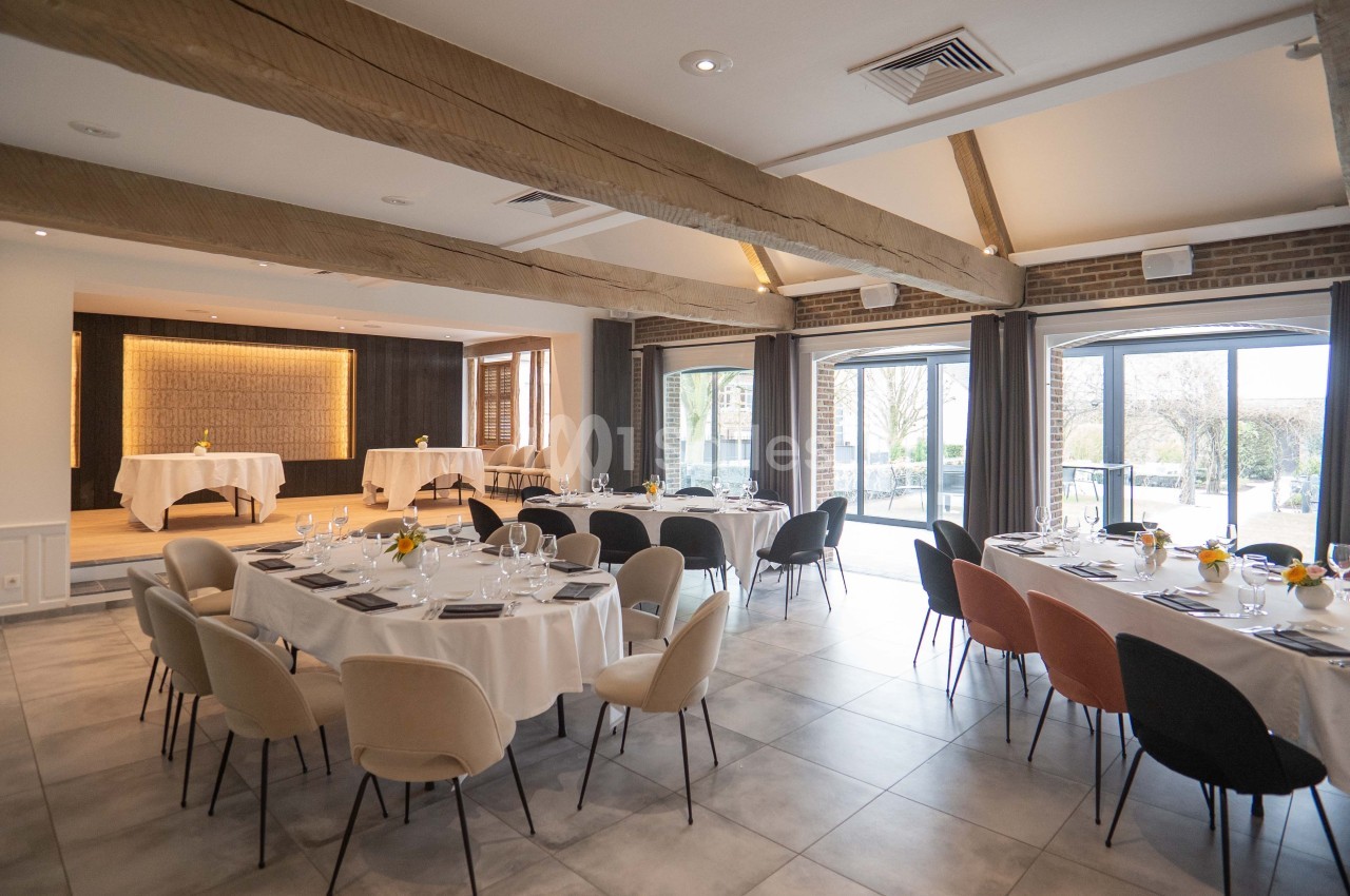 Salle lumineuse aménagée pour un événement, avec tables rondes dressées, chaises variées et grandes fenêtres donnant sur l…