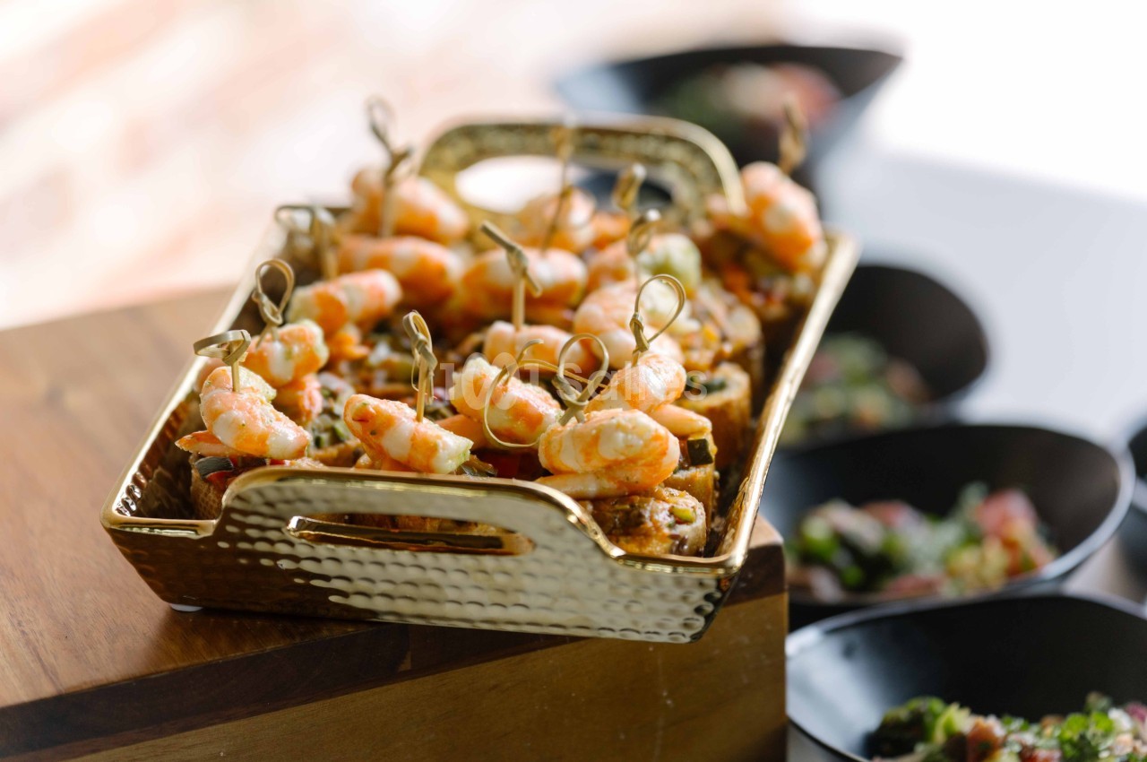 Plateau doré contenant des brochettes de crevettes servies sur des toasts, avec des bols en arrière-plan.