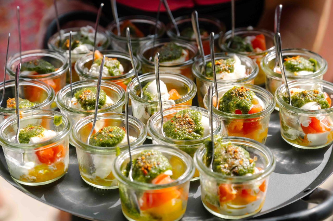 Plateau de verrines individuelles garnies de légumes, fromage et sauce verte, présentées avec des cuillères.
