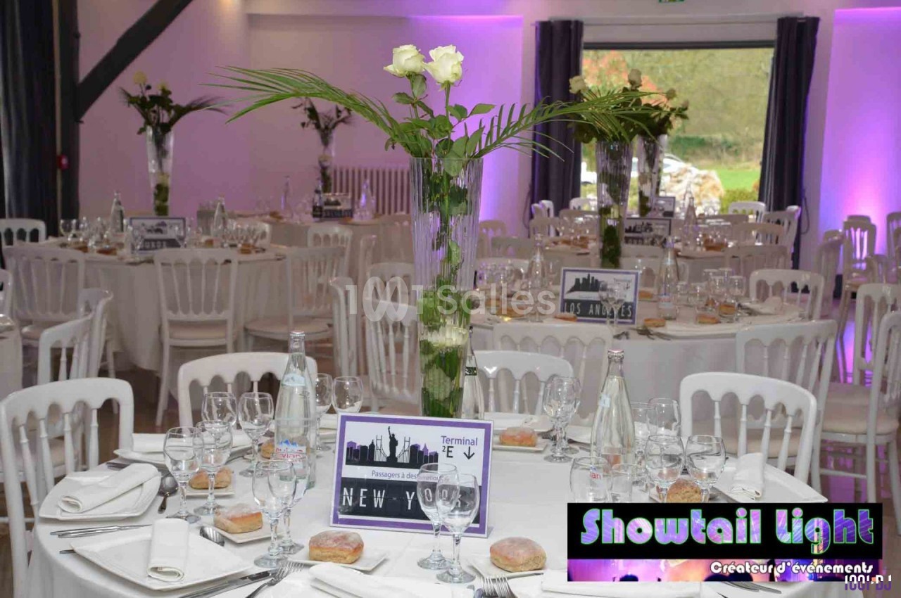Salle de réception décorée avec des tables rondes, nappes blanches, centres de table floraux et éclairage violet.