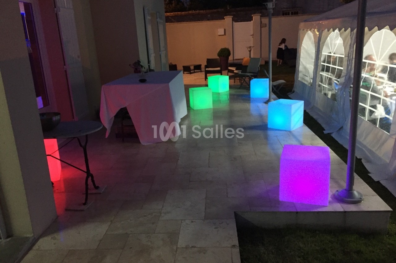 Cubes lumineux colorés disposés sur une terrasse en soirée, près d'une tente éclairée et d'une table dressée.