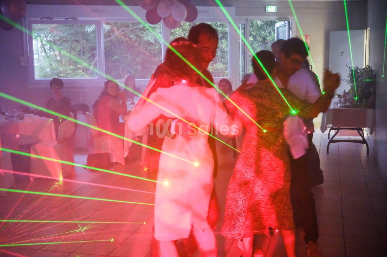 Des couples dansent sous des lumières laser colorées dans une salle éclairée par des fenêtres.