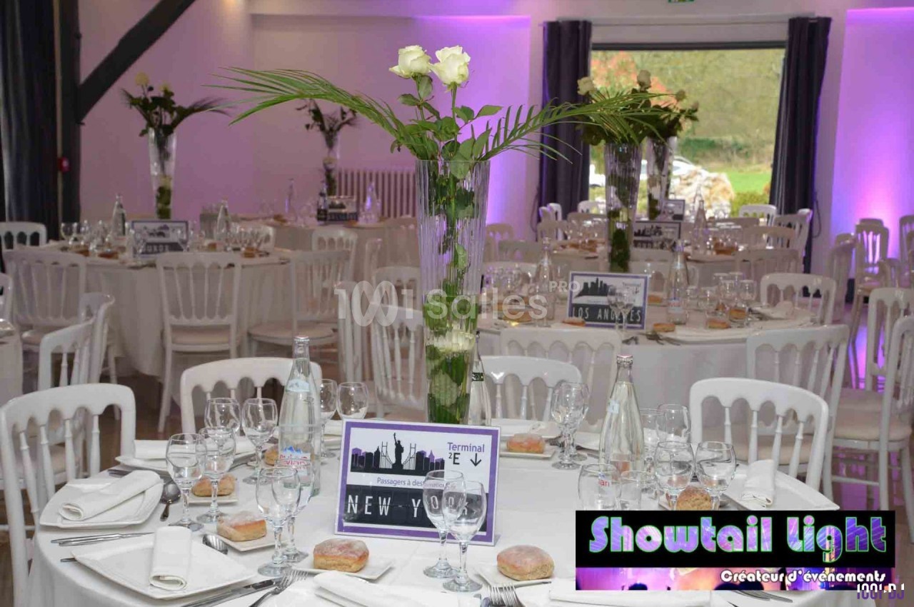 Salle de réception décorée avec des tables rondes, nappes blanches, centres de table floraux et éclairage violet.