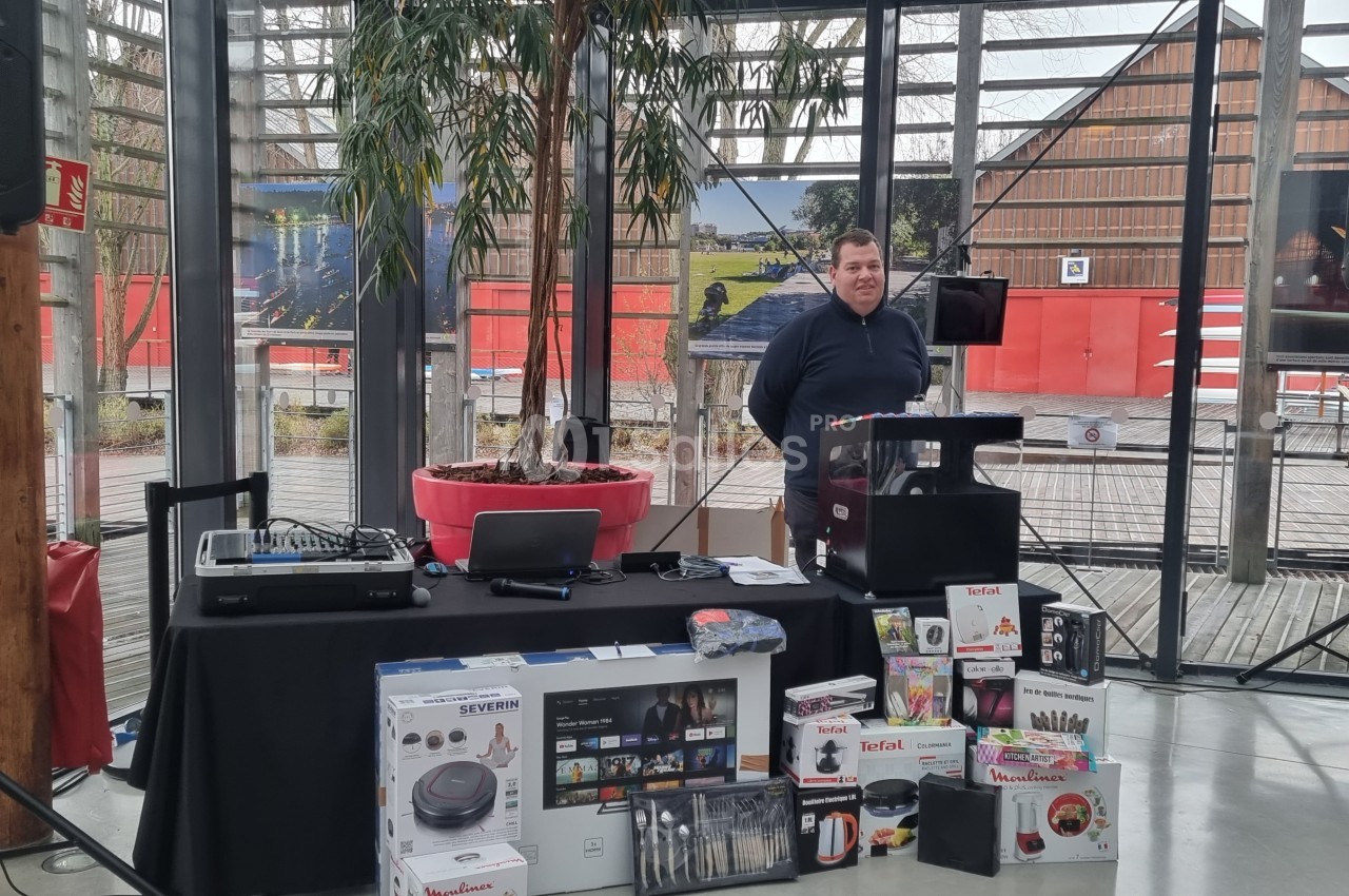 Un homme debout derrière un pupitre, près d'une table avec des lots exposés, dans un espace vitré.