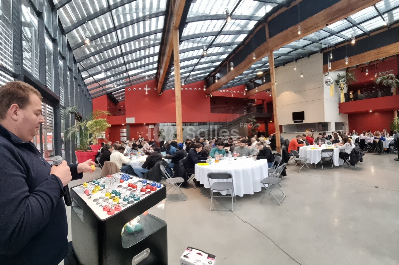 Un homme parle devant un public assis à des tables rondes dans une grande salle lumineuse avec verrière.