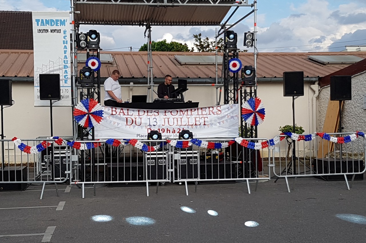 Scène extérieure décorée pour un bal des pompiers, avec deux personnes au stand DJ et des guirlandes tricolores.