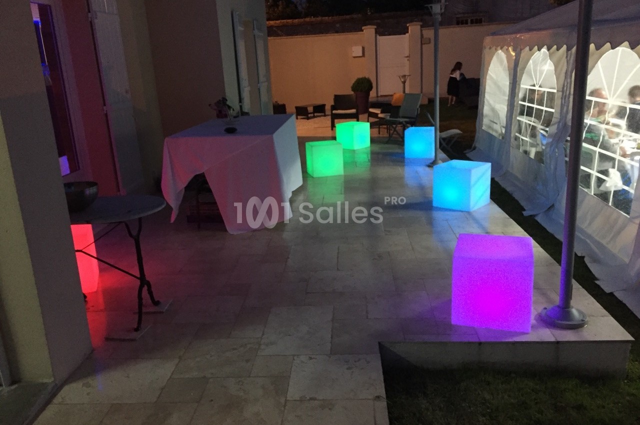 Cube lumineux coloré disposé sur une terrasse en soirée, près d'une tente blanche et d'une table dressée.