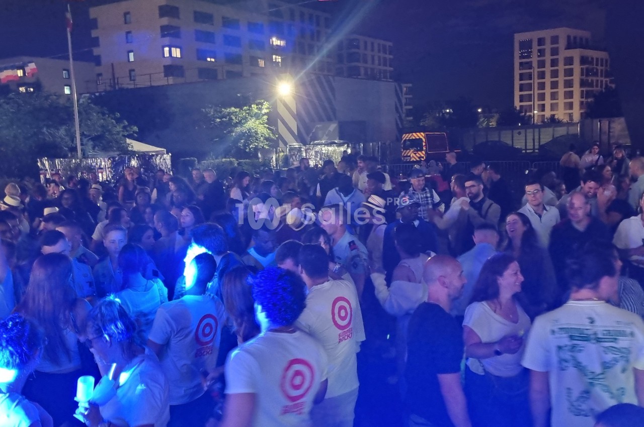 Foule dansante en extérieur de nuit, éclairée par des lumières colorées, avec des immeubles en arrière-plan.