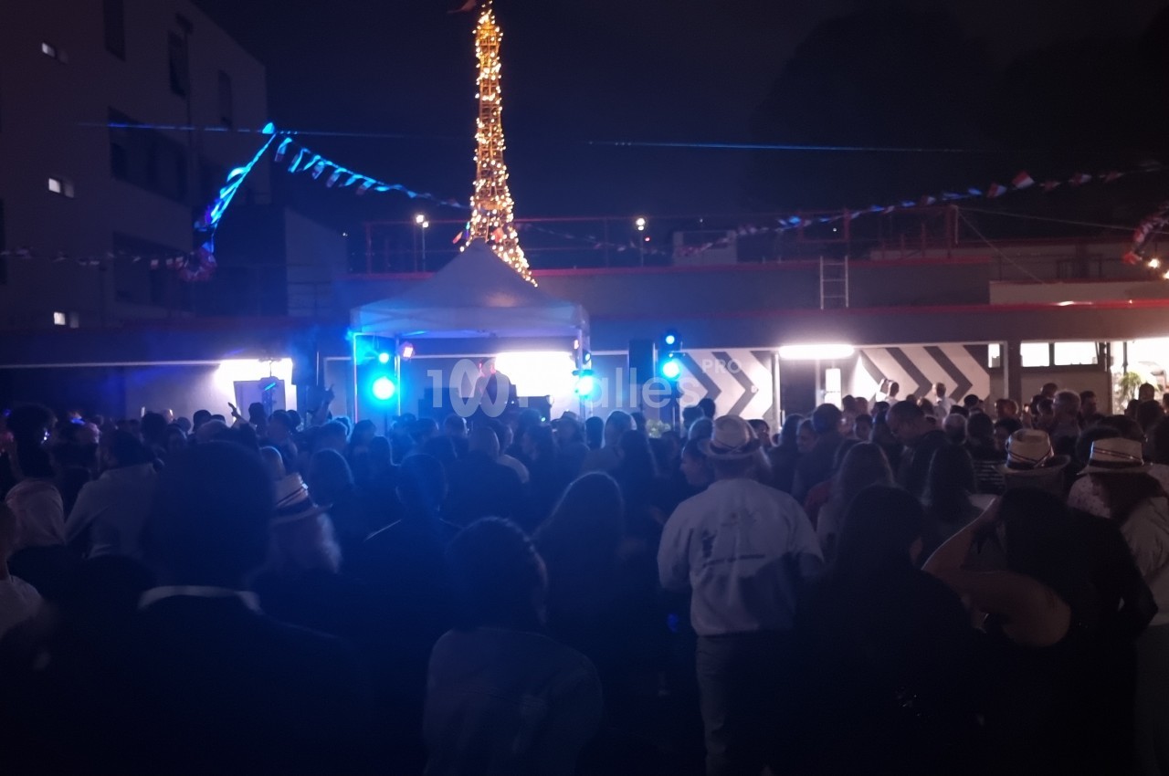 Foule rassemblée en soirée devant une scène éclairée, avec des décorations lumineuses en arrière-plan.