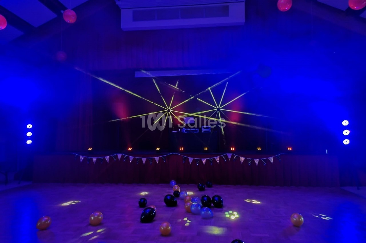 Salle de fête décorée avec des ballons au sol, éclairée par des lumières colorées et des lasers sur une scène.