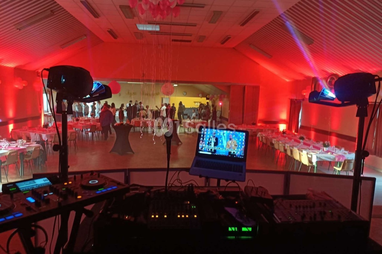 Salle de réception éclairée en rouge, vue depuis une table de mixage avec des invités et des tables dressées au fond.