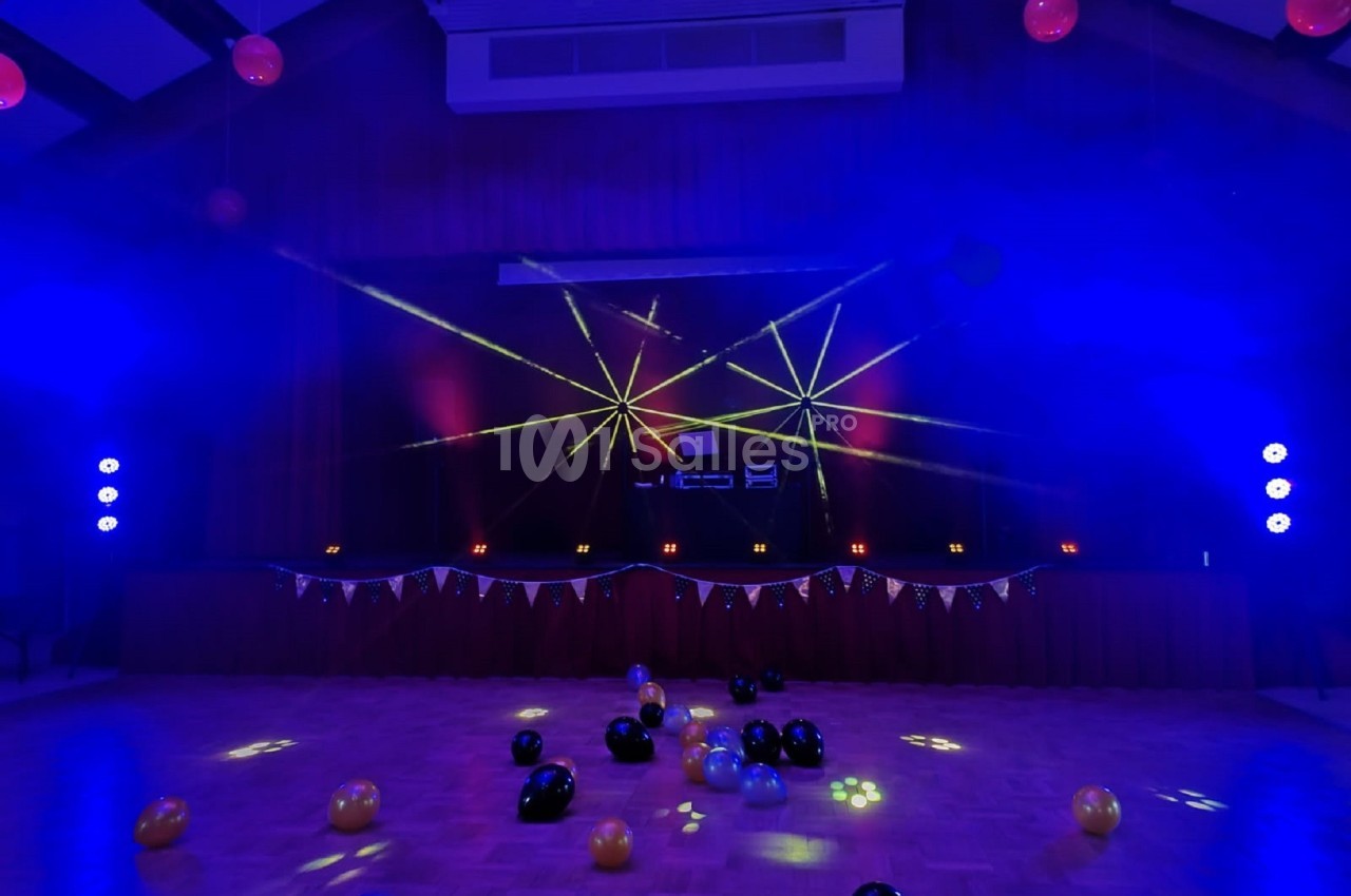 Salle de fête décorée avec des ballons au sol, éclairée par des lumières colorées et des lasers sur une scène.