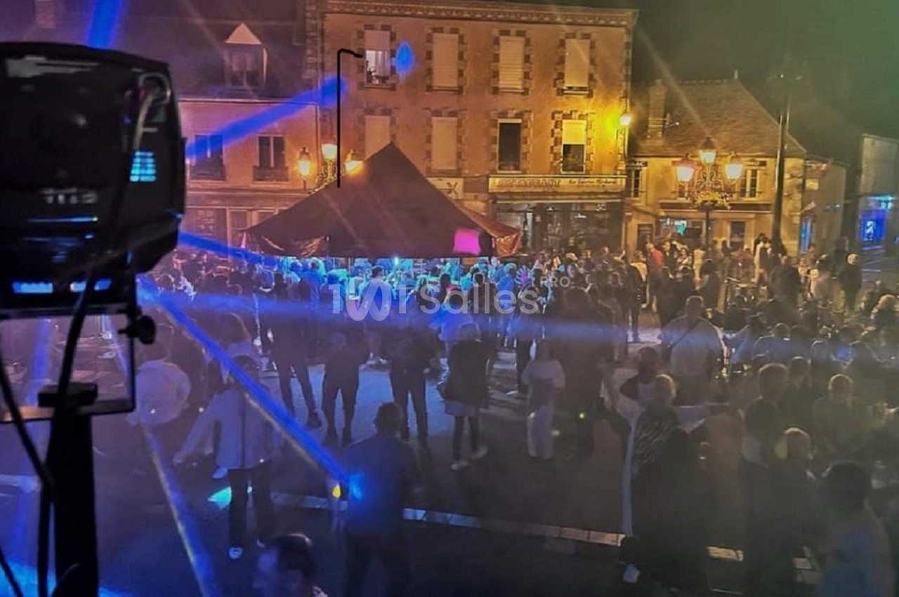 Foule rassemblée de nuit sur une place illuminée, avec des faisceaux lumineux et des bâtiments en arrière-plan.