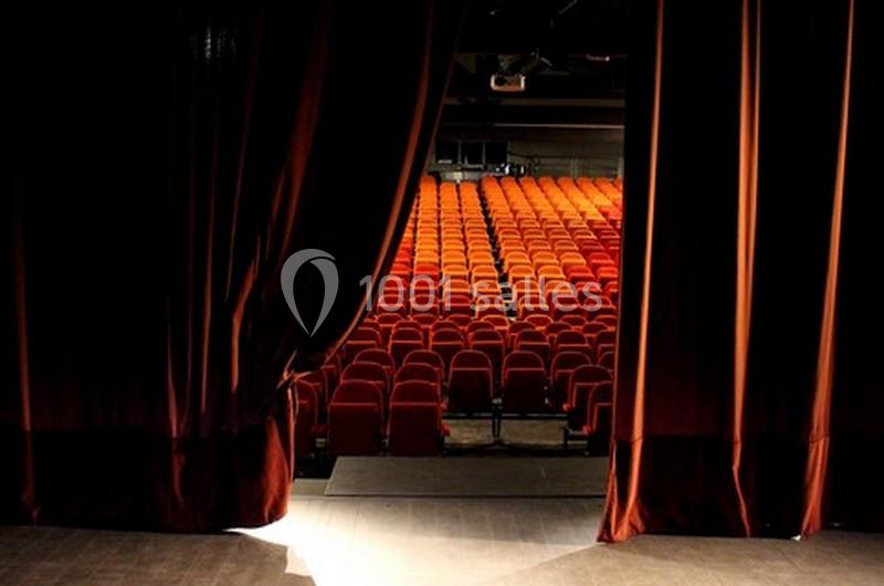 Vue depuis la scène d'une salle de théâtre avec des rideaux rouges ouverts et des rangées de sièges vides.