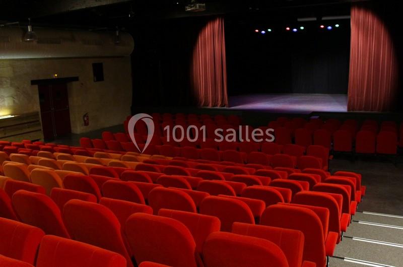 Salle de théâtre vide avec des rangées de sièges rouges et une scène éclairée par des projecteurs.