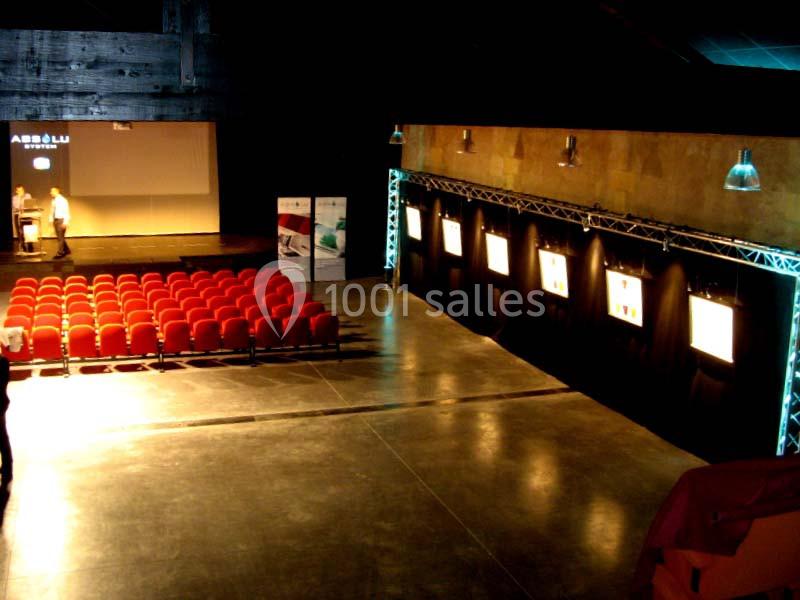 Salle d'exposition sombre avec rangées de sièges rouges, écrans lumineux sur un mur et projecteur en arrière-plan.