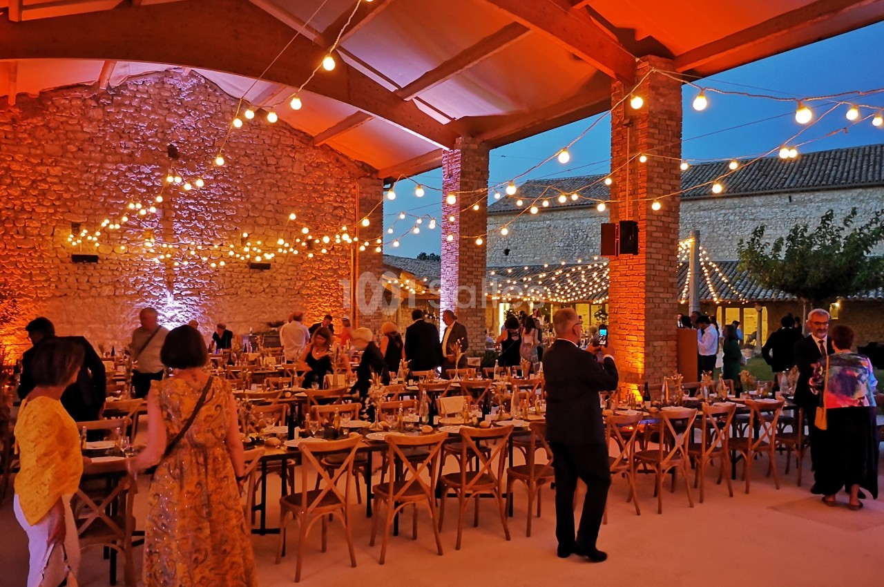 Salle de réception en soirée avec tables dressées, invités debout et guirlandes lumineuses suspendues.