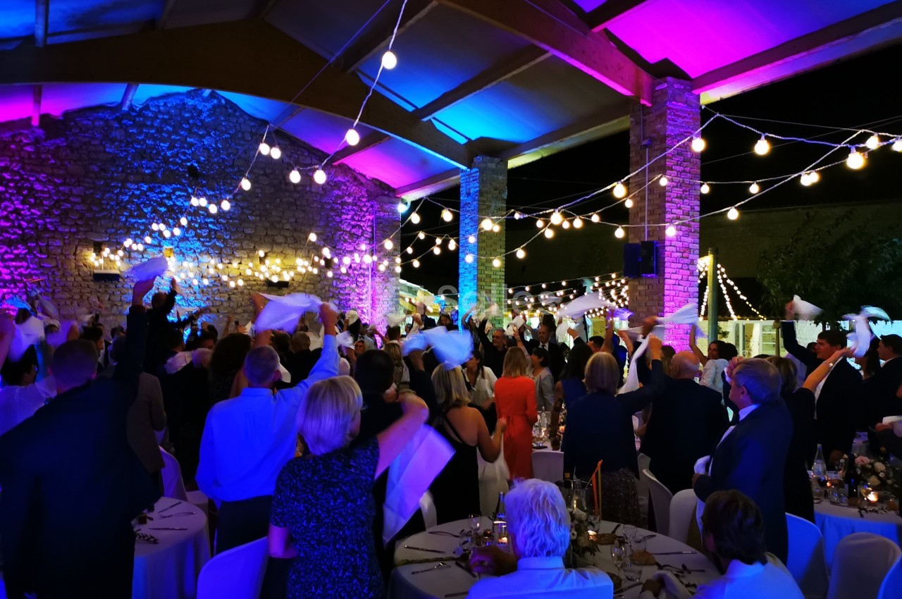 Des convives dans une salle illuminée agitent des serviettes blanches lors d'une fête ou d'un événement festif.