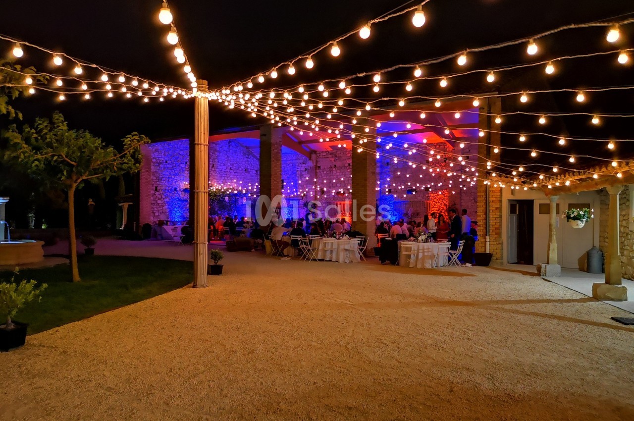 Cour extérieure illuminée par des guirlandes lumineuses, avec des tables dressées et des invités sous un éclairage coloré.