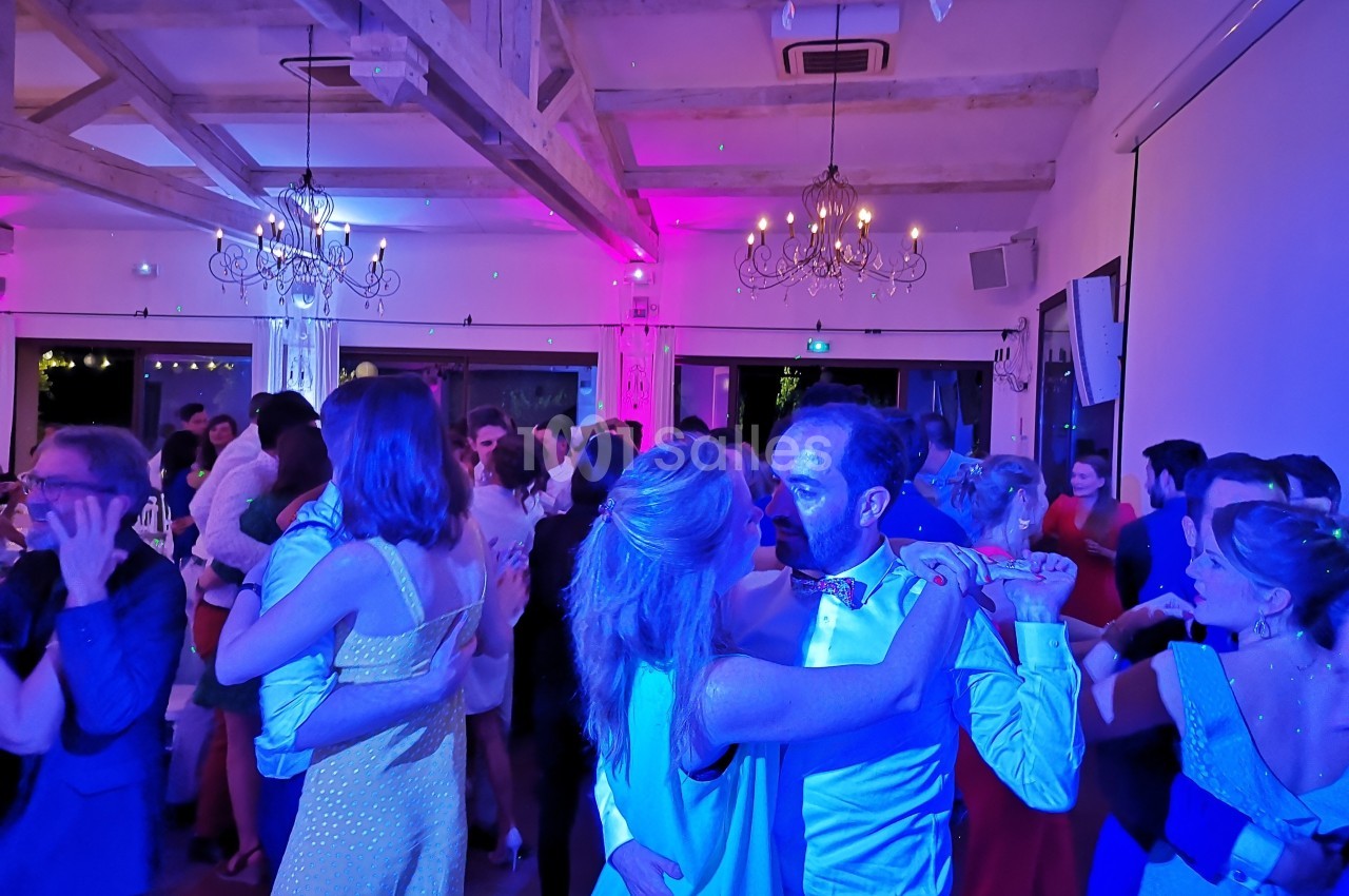 Des couples dansent dans une salle éclairée par des lumières colorées, avec des poutres apparentes et des lustres suspendus.